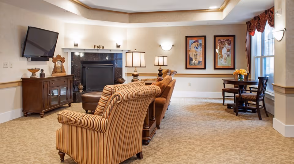 A cozy living room area with striped and solid upholstered armchairs arranged around a fireplace. A flat-screen TV is mounted on the wall above a wooden cabinet. Two framed artworks hang on the wall near a window with red valances. A small round table with two chairs and a vase of yellow flowers is positioned by the window. The room is warmly lit with wall sconces and table lamps.