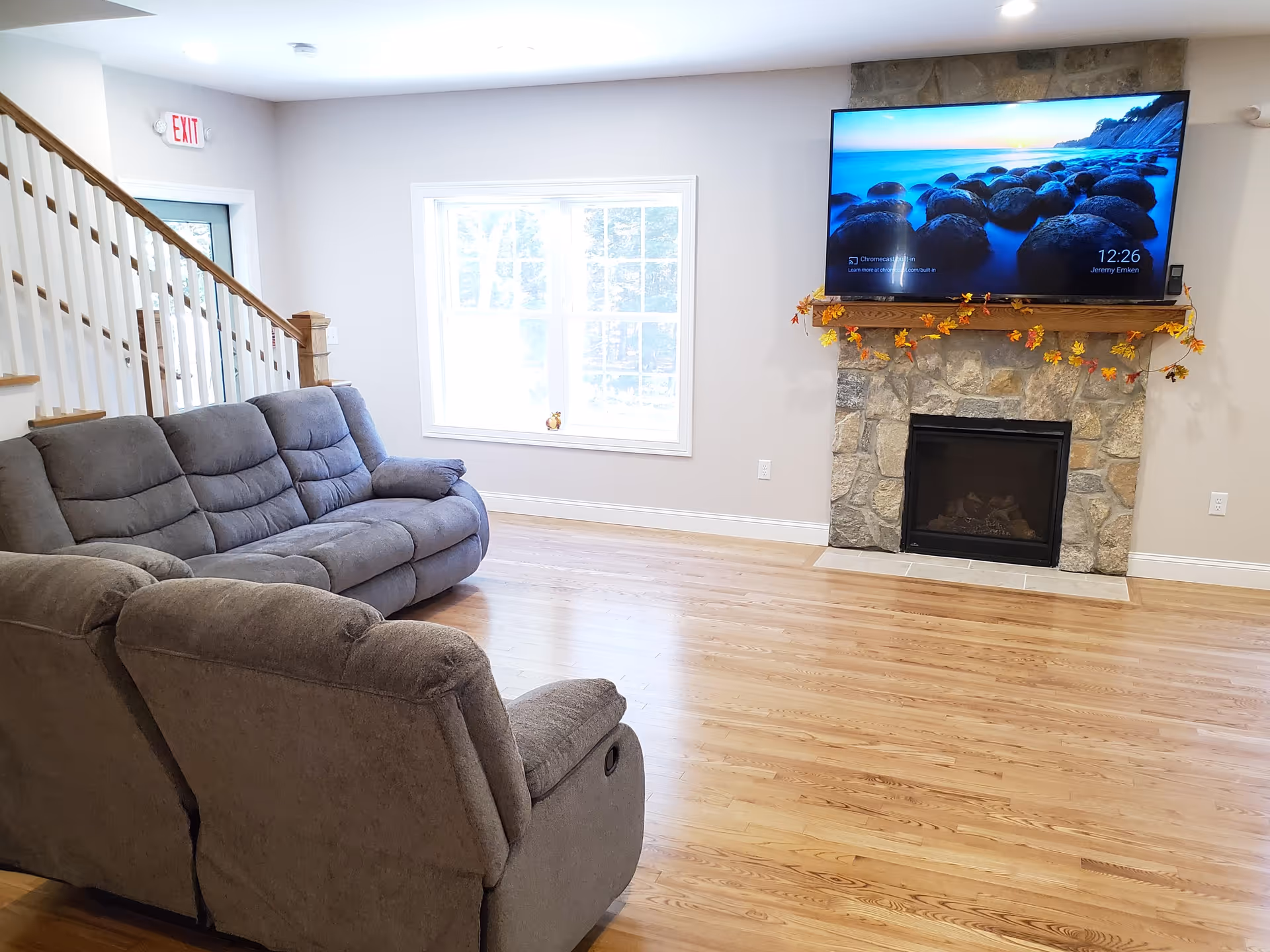 Bright living room with gray reclining sofas, hardwood floors, a stone fireplace with a mounted TV, and a staircase.