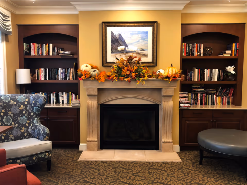 Cozy living room area with a stone fireplace decorated with autumn-themed garlands and pumpkins. On either side of the fireplace are built-in wooden bookshelves filled with books and decorative items. A floral patterned armchair is visible on the left and a round gray ottoman on the right. The walls are painted yellow and the floor is carpeted with a leaf pattern.