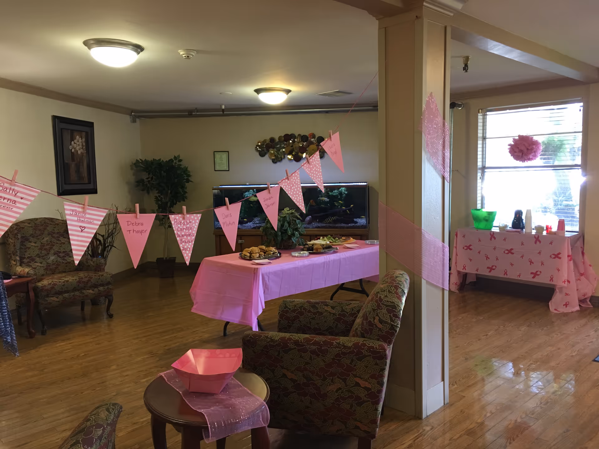 A decorated common area with wooden flooring and beige walls. There are two upholstered chairs and a small round table with a pink bowl and pink cloth. Pink triangular pennant banners with handwritten names hang across the room. A table covered with a pink tablecloth holds plates of sandwiches and snacks. In the background, there is a large fish tank and a table near a window decorated with pink ribbons and breast cancer awareness symbols. The room is softly lit with ceiling lights.