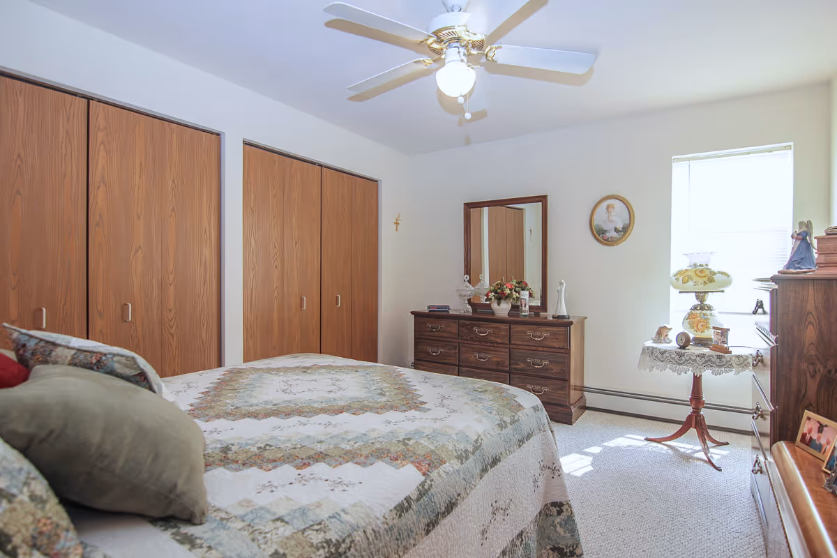 A cozy bedroom with a large bed covered in a patterned quilt. The room features wooden closets with sliding doors, a wooden dresser with a mirror, and a small round table by the window holding decorative items and a vintage lamp. A ceiling fan with a light is mounted on the white ceiling, and the room is softly lit by natural light coming through the window.