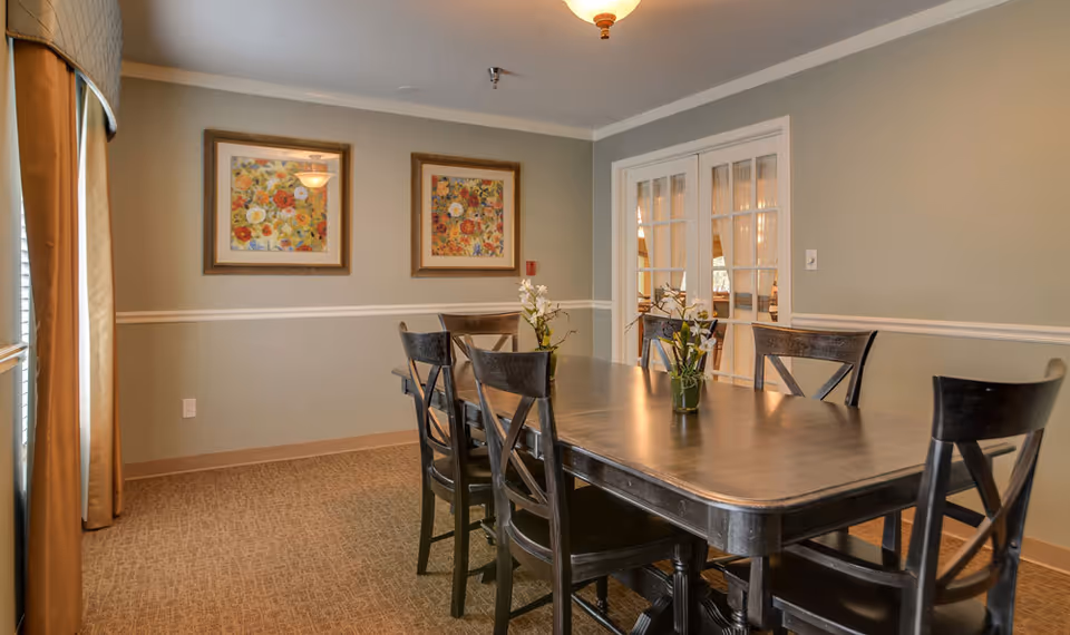 A dining room with a long wooden table surrounded by six dark wooden chairs. Two small vases with flowers are placed on the table. The room has beige walls with white trim, two framed floral paintings on the wall, and a window with beige curtains on the left side. French doors with glass panes are visible in the background.