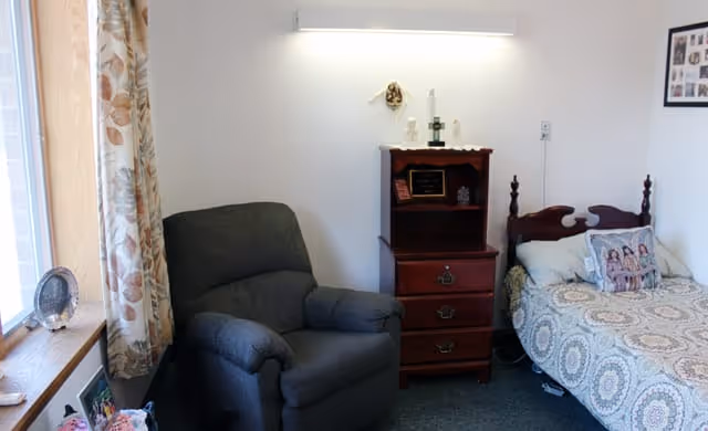 A cozy bedroom featuring a single bed with patterned bedding and a decorative pillow, a dark upholstered armchair, a wooden nightstand with drawers and decorative items on top, and a window with floral curtains letting in natural light.