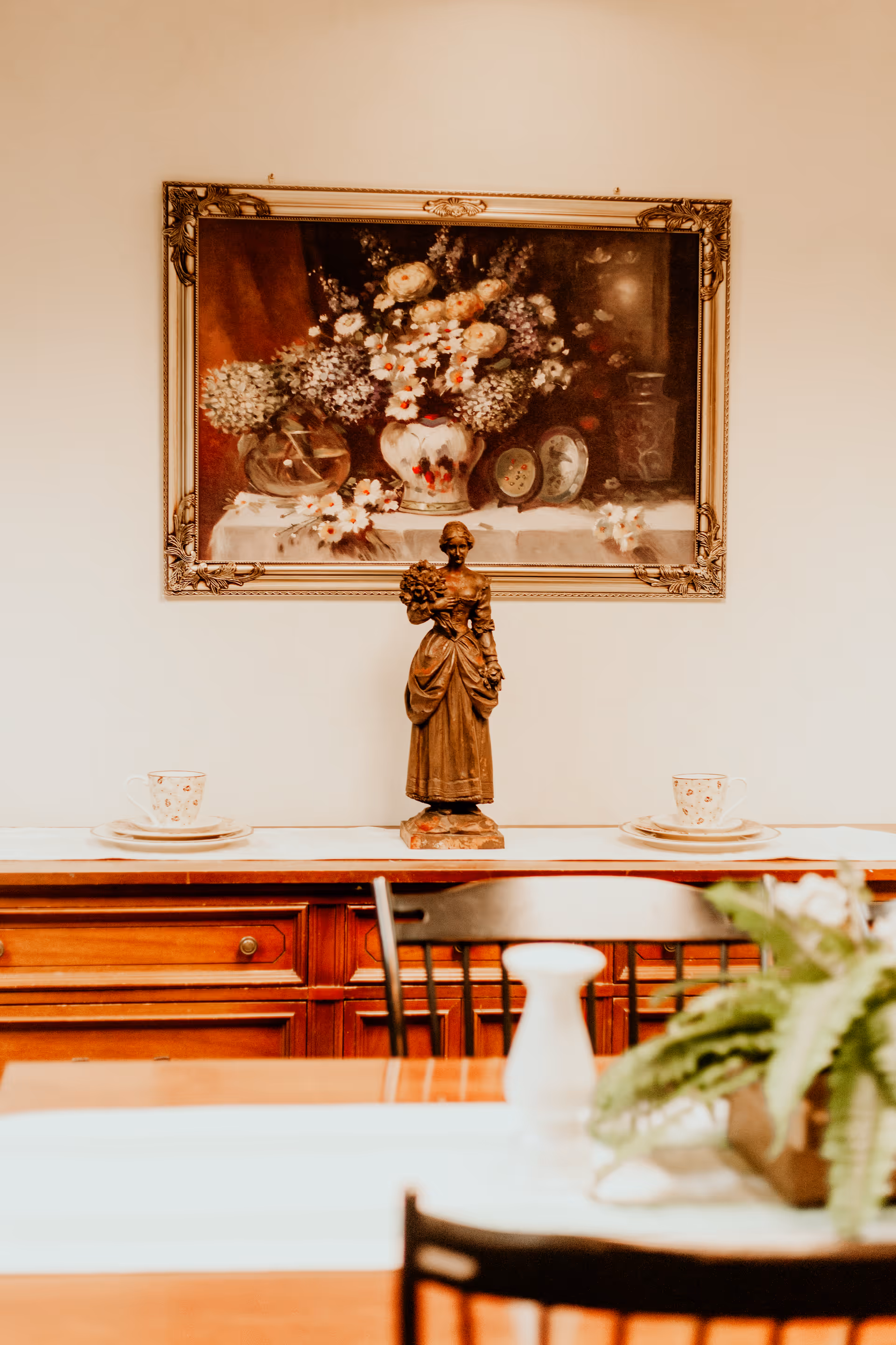A wooden sideboard with two teacups and saucers placed on top, a bronze statue of a woman holding flowers in the center, and a framed painting of a floral arrangement hanging on the wall above.