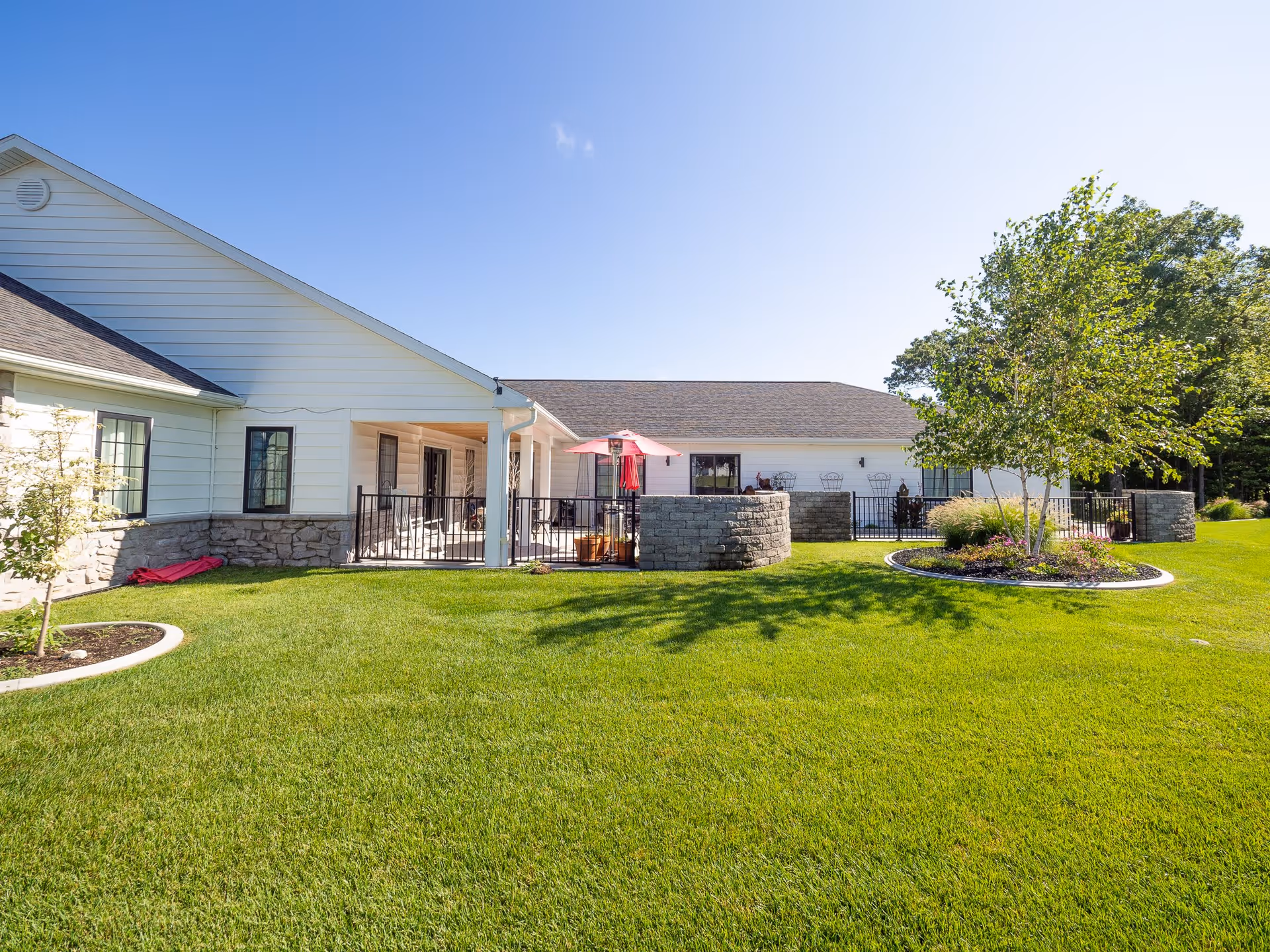 Exterior view of a single-story senior care facility building with white siding and stone accents. The building has a covered patio area with a red umbrella and outdoor seating. The foreground features a well-maintained green lawn with landscaped flower beds and trees under a clear blue sky.