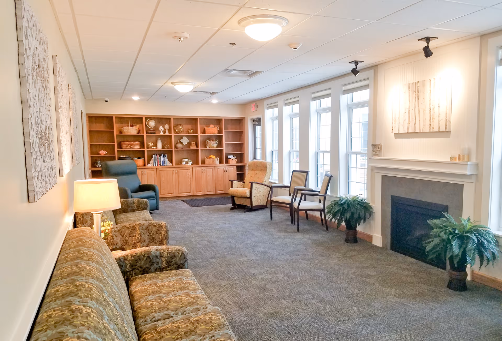 A bright and cozy living room area in Birchwood Cottages - Owatonna featuring a patterned sofa, armchairs, a wooden bookshelf filled with decorative items and books, large windows letting in natural light, a fireplace with plants on either side, and wall art above the fireplace.