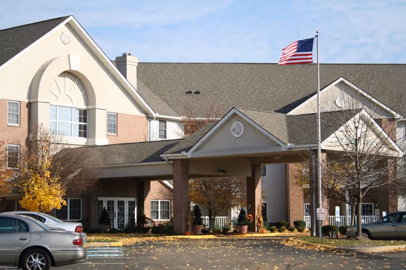 Exterior view of a senior living facility building with a covered entrance, an American flag on a flagpole, several parked cars, and trees with autumn foliage under a partly cloudy sky.