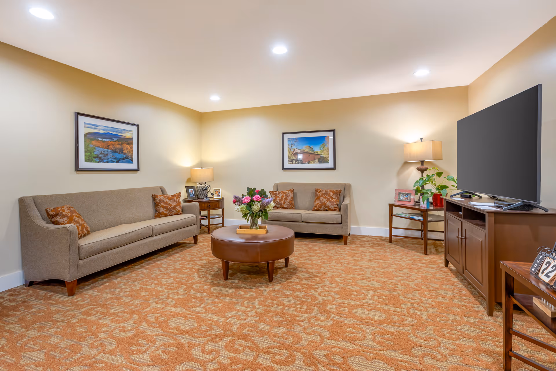 A cozy living room with two beige sofas adorned with patterned orange pillows, a round brown ottoman with a vase of flowers on top, two side tables with lamps and framed photos, a large flat-screen TV on a wooden cabinet, and two framed landscape pictures on the beige walls. The floor is covered with an orange patterned carpet.