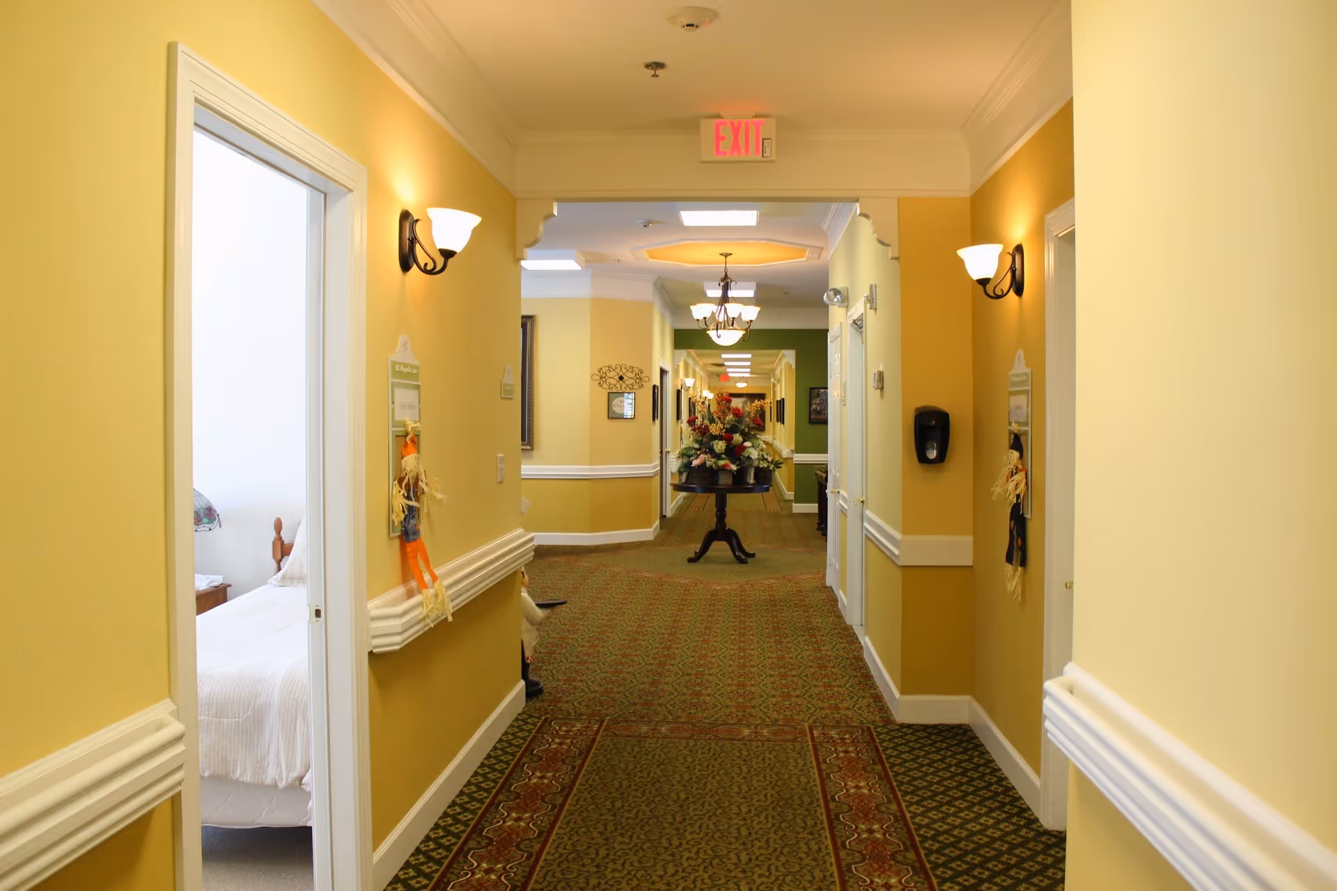 A well-lit hallway in a senior living facility with yellow walls, wall sconces, and patterned carpet. Doors line both sides of the hallway, one of which reveals a bedroom with a bed and nightstand. At the end of the hallway, there is a table with a large floral arrangement. An exit sign is visible on the ceiling.