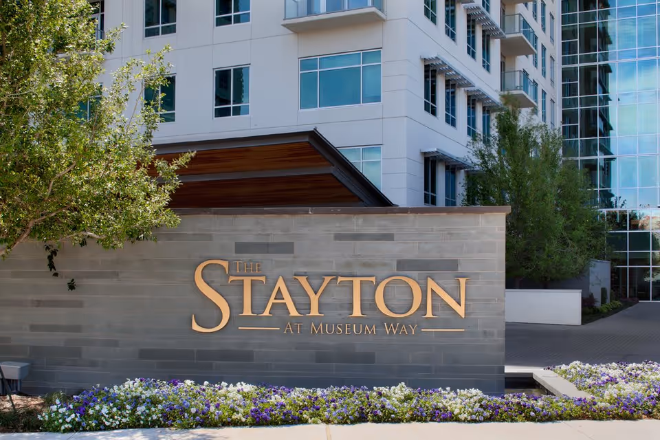 Exterior view of The Stayton at Museum Way sign mounted on a gray stone wall with a flower bed of white and purple flowers in front. Behind the sign is a modern multi-story building with large windows and some trees.