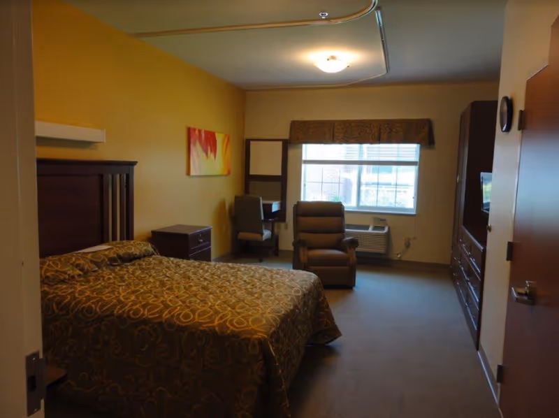 A bedroom in Woodbury Wellness Center featuring a bed with a patterned bedspread, a nightstand, a recliner chair, a desk with a chair, a window with blinds and a valance, an air conditioning unit below the window, and a dresser with a mounted TV.