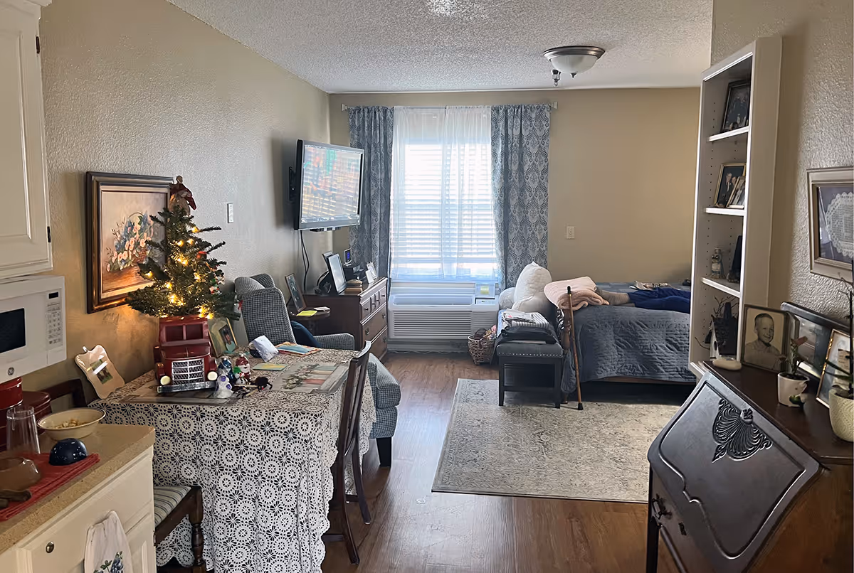 Interior view of a senior living facility room at Broadmoor Court showing a cozy living and sleeping area. The room features a small kitchen counter with a microwave and bowls, a table covered with a lace tablecloth decorated with a small lit Christmas tree and various items. There is a comfortable armchair facing a wall-mounted TV, a window with patterned curtains, and a bed with a gray quilt and a walking cane resting beside it. Shelves with framed photos and decorations are visible, along with a wooden desk and a rug on the wooden floor.