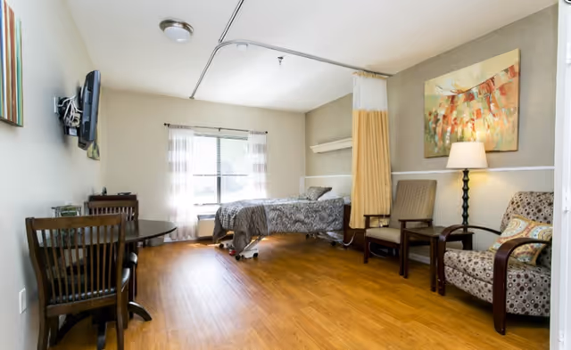 A well-lit senior living facility room with a single bed near a window covered with sheer curtains. The room has wooden flooring, a round table with chairs, two armchairs, a floor lamp, and a colorful abstract painting on the wall. A privacy curtain is partially drawn near the bed.