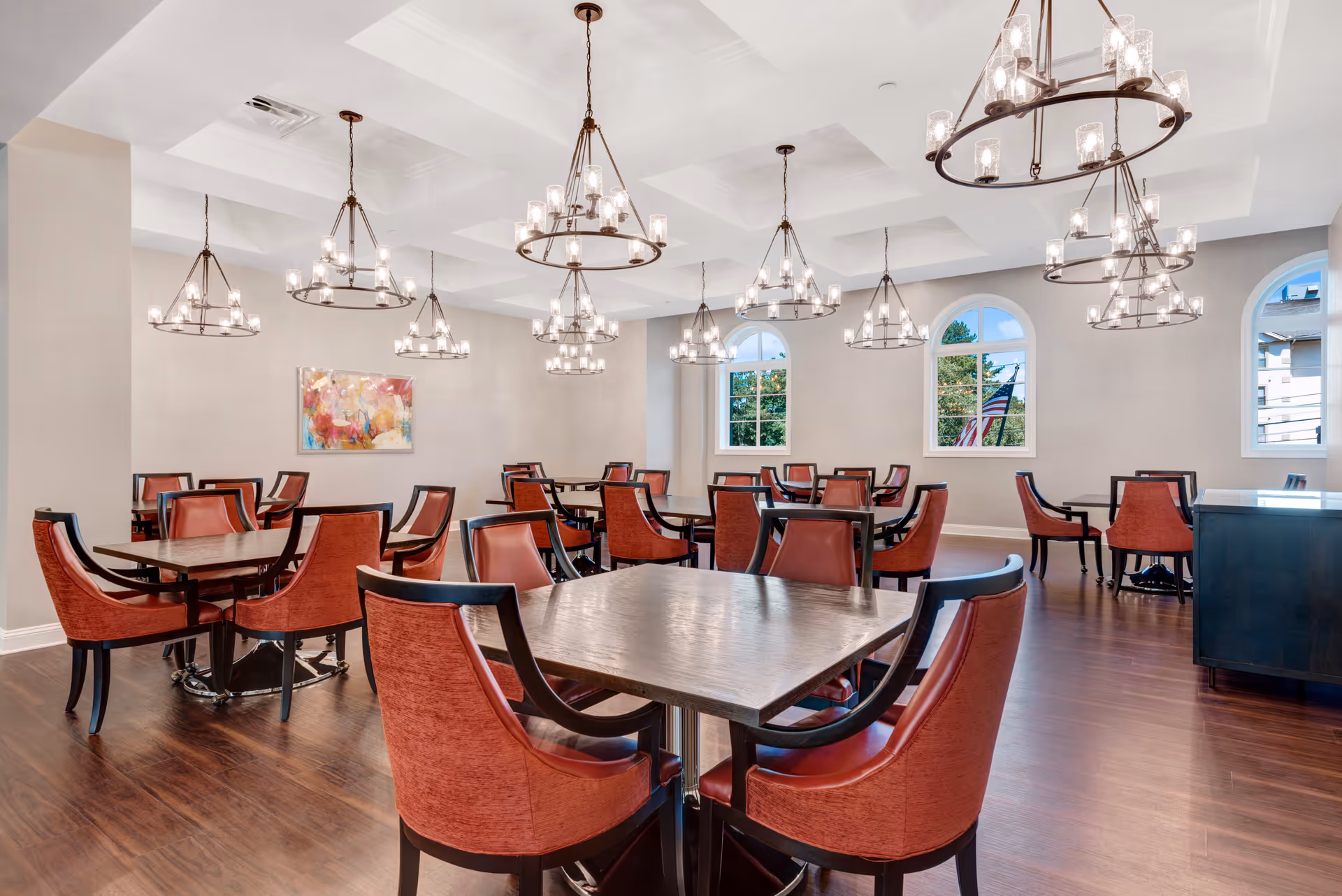 A spacious dining room with multiple square wooden tables surrounded by red upholstered chairs. The room features large arched windows letting in natural light, several modern circular chandeliers hanging from a coffered ceiling, and a colorful abstract painting on one wall. The floor is a polished dark wood.