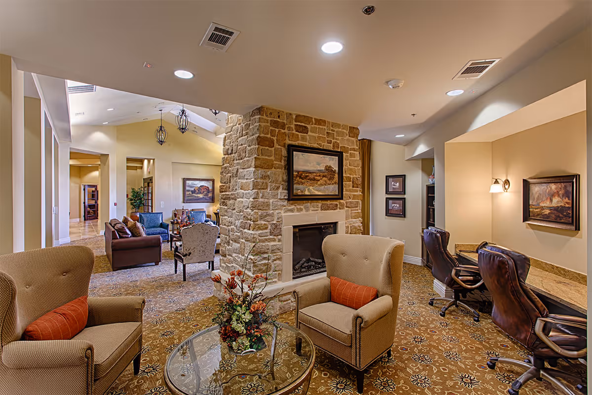 A cozy and well-lit common area in a senior living facility featuring a stone fireplace with a framed painting above it. The room has comfortable armchairs with orange cushions, a glass coffee table with a floral arrangement, and a carpet with a decorative pattern. In the background, there are additional seating areas with sofas and chairs, as well as a workspace with two leather office chairs and a granite countertop. The walls are painted in warm beige tones and decorated with framed artwork.