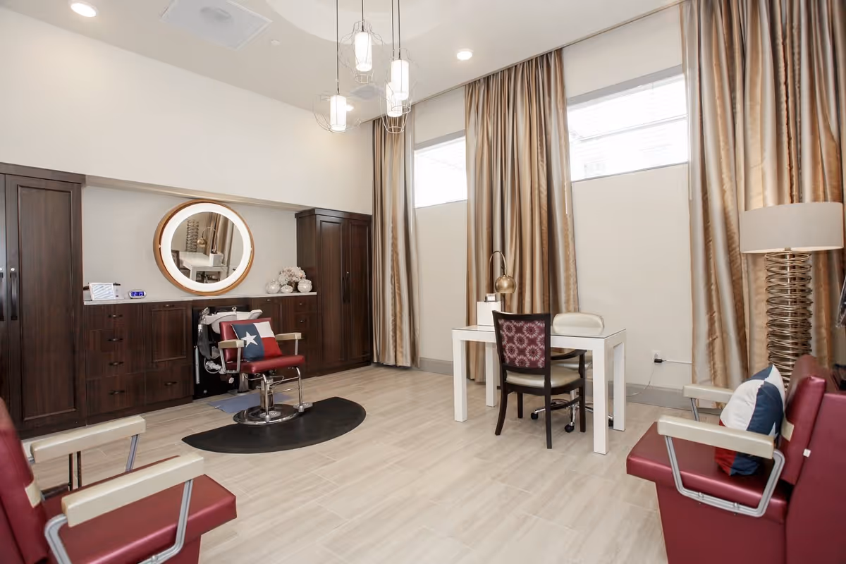 A well-lit room with beige tiled flooring and cream-colored walls featuring tall windows with gold curtains. The room contains a salon chair with a star-patterned pillow in front of a round mirror mounted on a dark wooden cabinet. There are two maroon chairs with armrests and pillows, a white desk with two chairs, and a modern floor lamp with a spiral design. Three pendant lights hang from the ceiling.