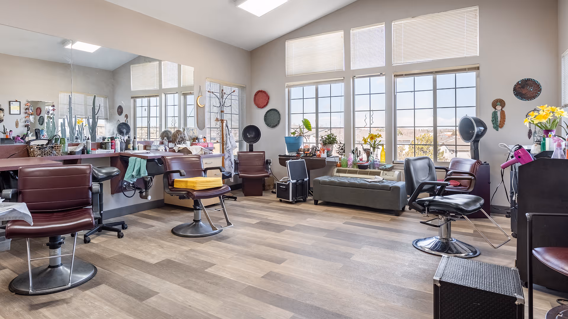 A bright and spacious hair salon area with multiple styling chairs, large mirrors, and various hair care products on the counters. The room has large windows letting in natural light, plants on the windowsill, and hair drying equipment. The floor is wooden and the walls are decorated with small art pieces.