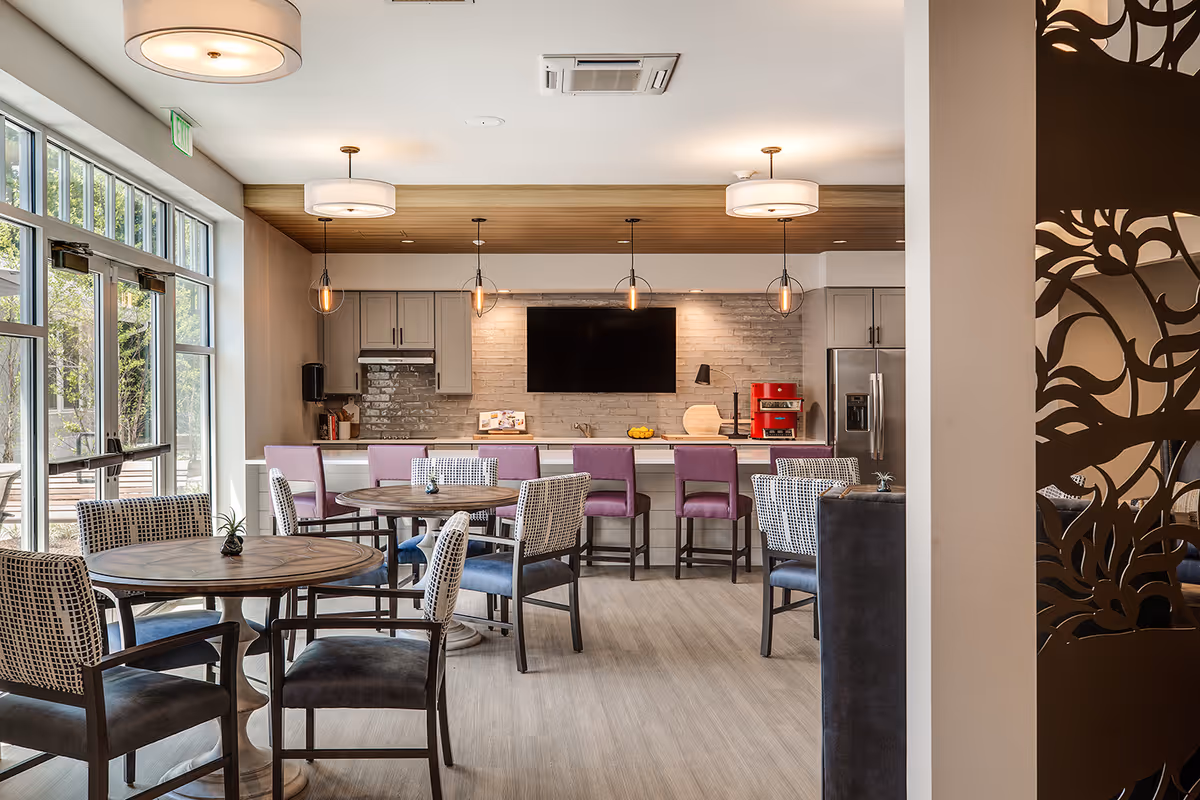 A bright and modern communal dining area with round wooden tables and cushioned chairs. The room features large windows letting in natural light, a kitchen area with a countertop and bar stools, pendant lights hanging from the ceiling, a wall-mounted TV, and a red coffee machine on the counter. Decorative elements include a patterned room divider on the right side.