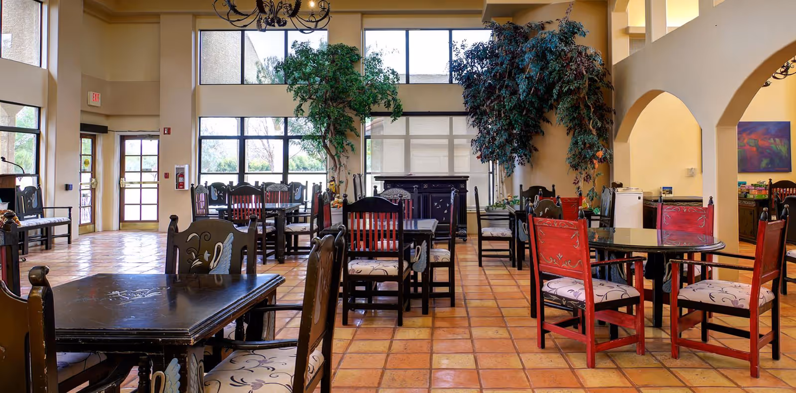 Spacious dining area with multiple wooden tables and chairs, tiled floor, large windows, and potted trees.