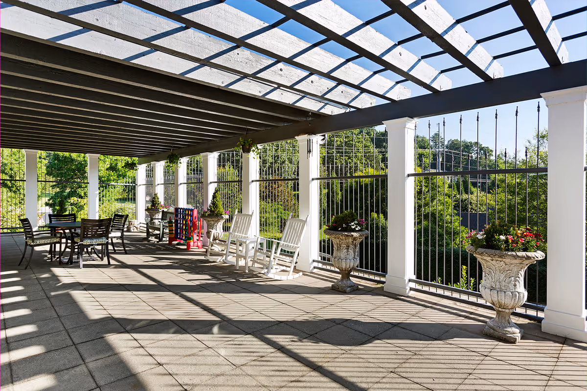Shaded outdoor terrace with a pergola, white columns, rocking chairs, tables, and decorative planters overlooking green trees.
