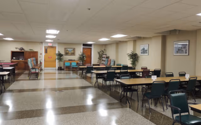 A spacious room with multiple rectangular tables and chairs arranged in rows, likely used for dining or group activities. The room has a polished floor with a pattern, beige walls adorned with framed pictures, and several potted plants. There are doors and a wooden cabinet visible in the background under fluorescent ceiling lights.