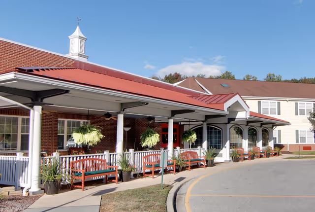Exterior view of a senior living facility named Dover Place featuring a covered porch with red benches, hanging plants, and potted plants along the walkway. The building has a red roof and brick and light-colored siding walls under a clear blue sky.