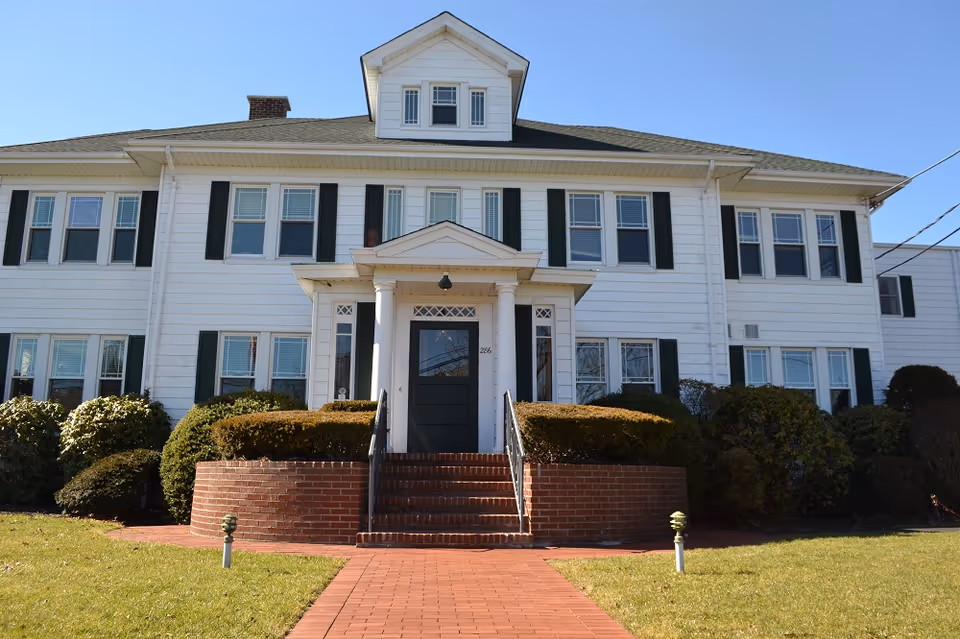Front exterior view of a two-story white building with black shutters, a central entrance with a small porch supported by columns, brick steps, and a well-maintained lawn and bushes in front under a clear blue sky.