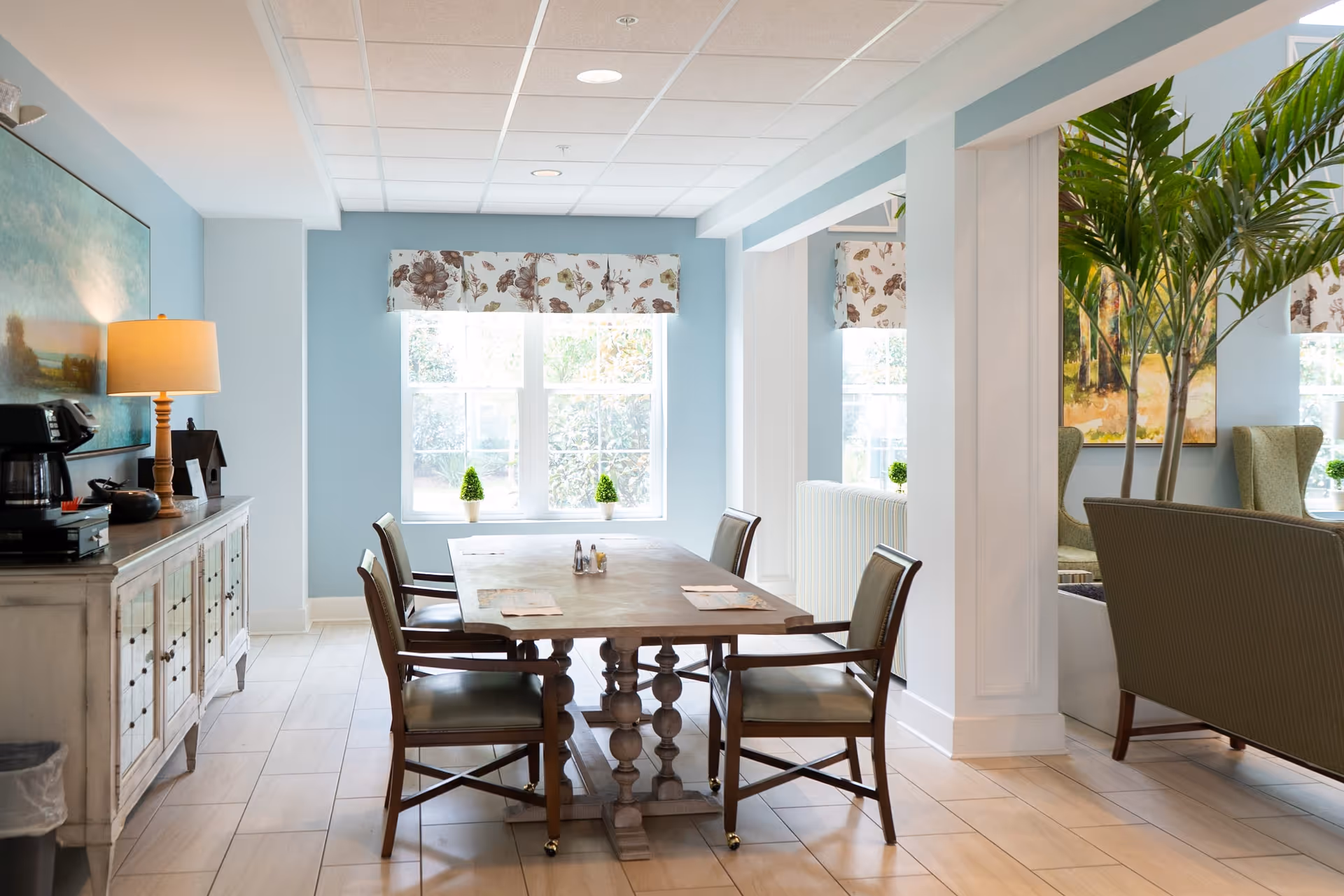 A bright dining area with a wooden table and four chairs. The room has light blue walls, a large window with floral valances, and a sideboard with a lamp and coffee maker. There are small potted plants on the windowsill and a large green plant near a seating area with armchairs.