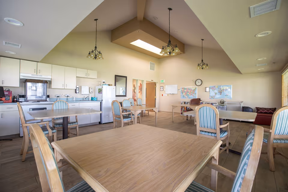 A spacious dining area with multiple wooden tables and chairs with striped cushions. The room features a kitchenette with white cabinets, a stove, refrigerator, and various kitchen appliances. The ceiling has a skylight and hanging light fixtures. Maps and a clock are mounted on the beige walls, and large windows allow natural light to fill the room.