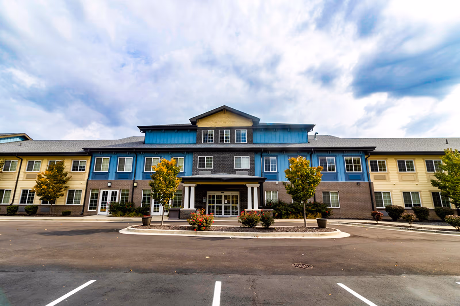 Front exterior view of a multi-story senior living facility building with a covered entrance, several windows, small trees, and landscaped bushes under a cloudy sky.