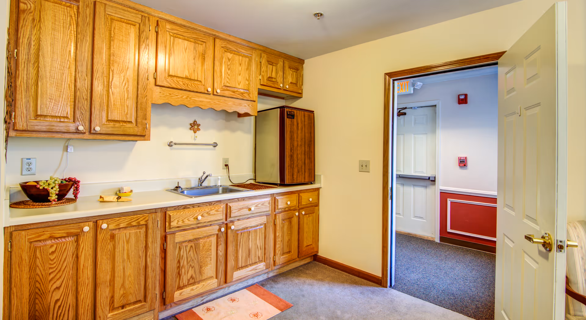 Interior view of a small kitchen area with wooden cabinets, a countertop with a sink, a small refrigerator, and a bowl of fruit on the counter. The room has beige walls and carpeted floor. An open door leads to a hallway with white walls and red wainscoting.
