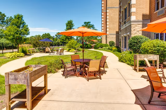 Sunlit outdoor courtyard with round tables, orange umbrellas, chairs, a paved walkway, and landscaped greenery beside a brick senior living building.