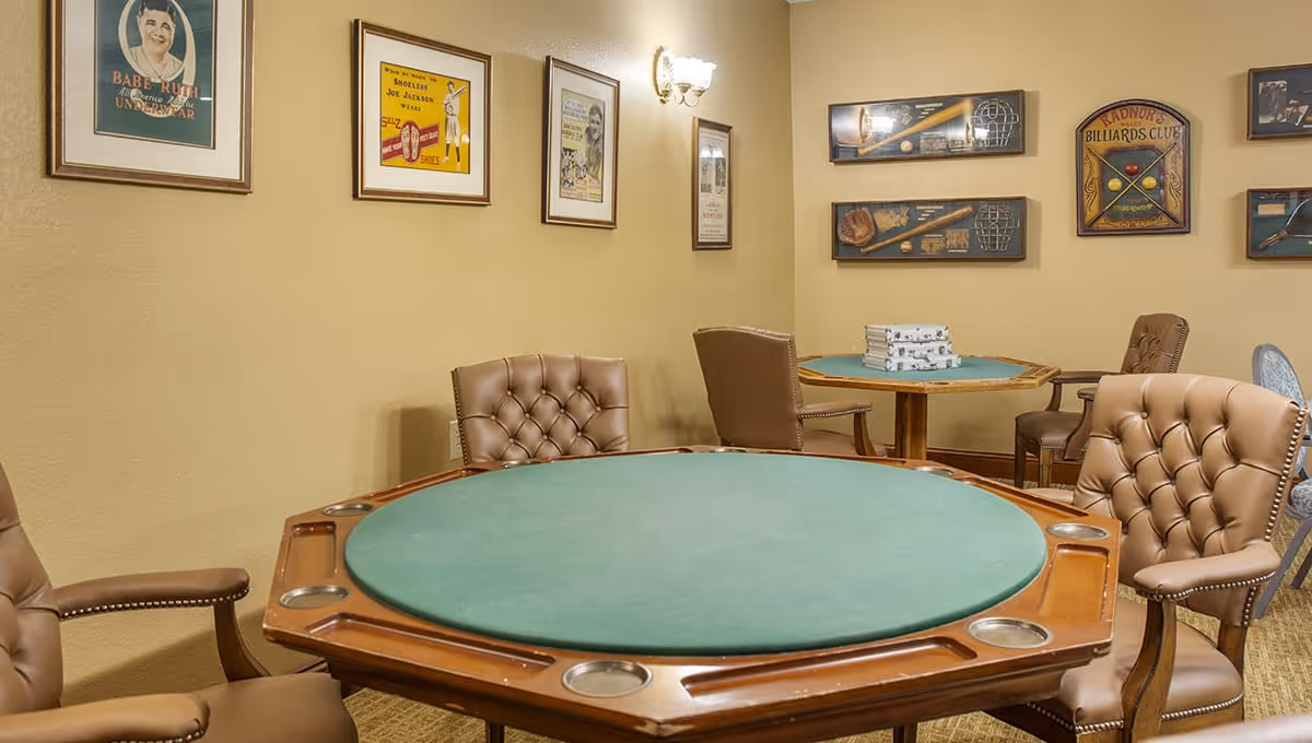 A game room with a large round poker table covered in green felt surrounded by four brown leather tufted chairs. In the background, there is another smaller round table with chairs and a stack of board games on it. The walls are decorated with framed vintage sports posters and billiards-themed artwork.