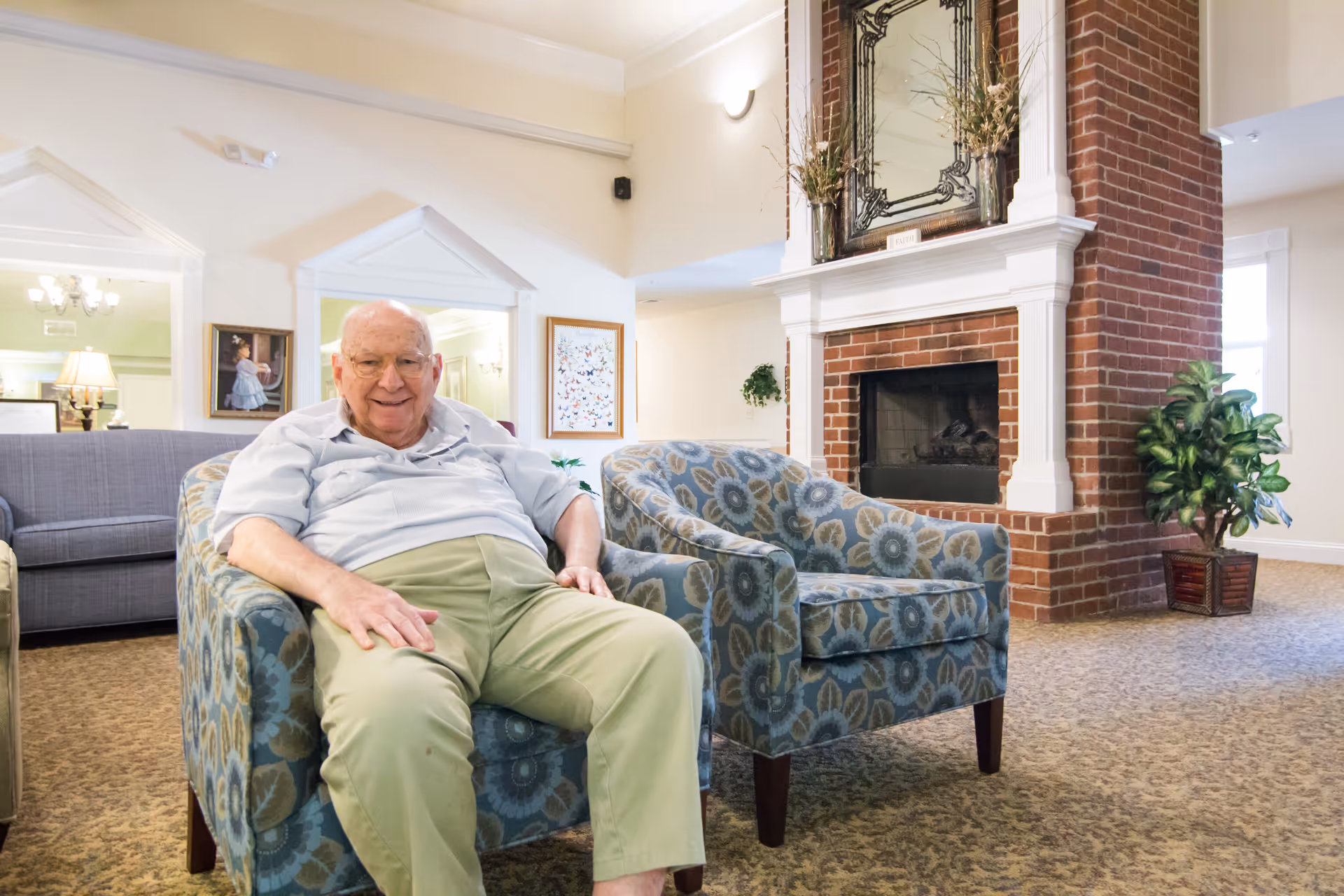 An elderly man sitting and smiling on a patterned armchair in a cozy living room with a brick fireplace, decorative mirror above the mantel, another matching armchair, a gray sofa, framed artwork on the walls, and a potted plant in the corner.