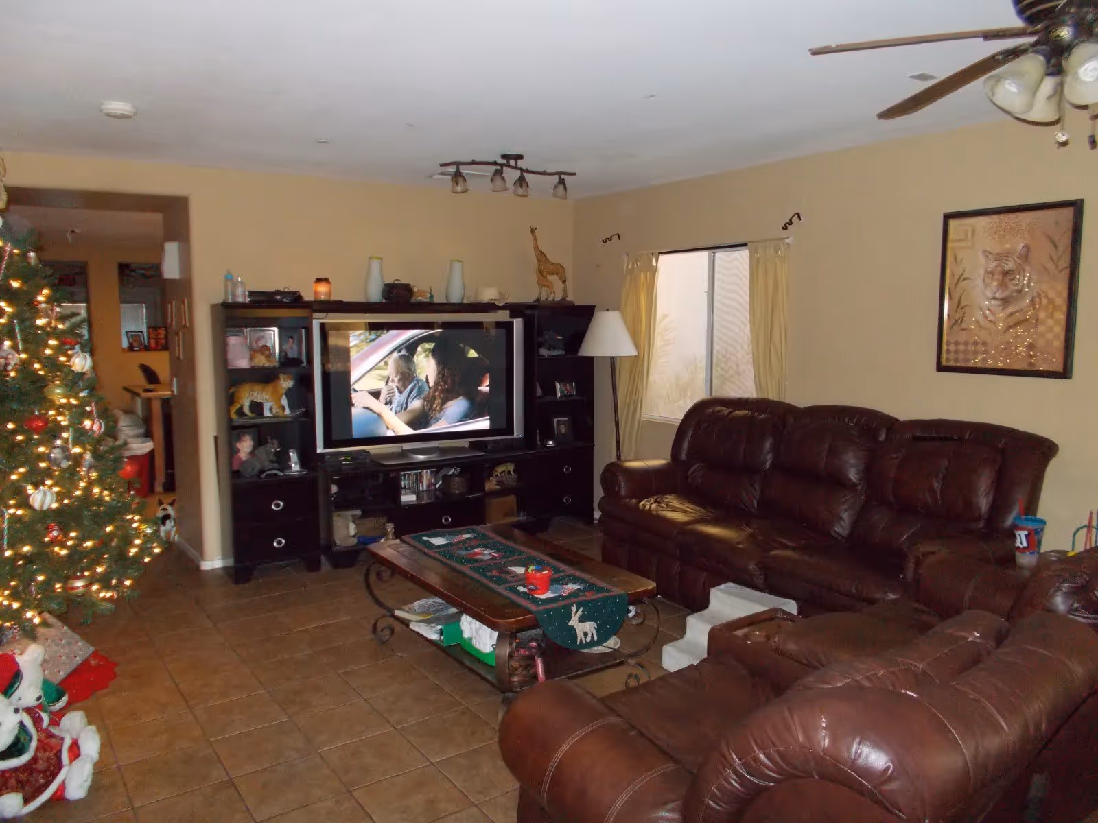 A cozy living room with brown leather sofas, a wooden coffee table decorated with a Christmas-themed runner, a decorated Christmas tree with lights on the left, a large TV on a black entertainment center showing a scene with two people in a car, a window with beige curtains, a ceiling fan with lights, and a framed picture of a tiger on the wall.