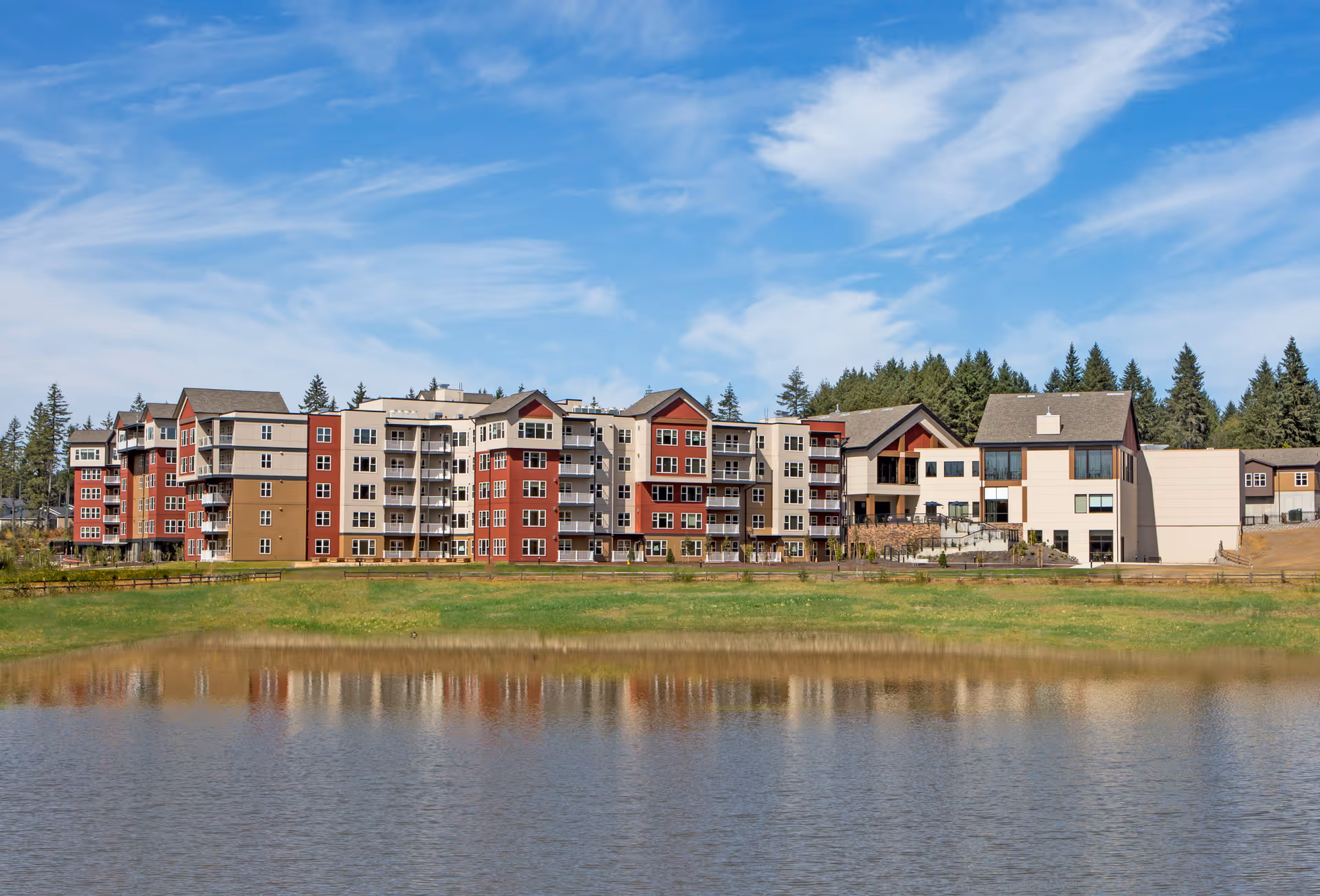 Wide exterior view of a multi-story senior living facility named Heron's Key, featuring a combination of red, beige, and brown buildings with balconies, set against a backdrop of trees and a partly cloudy blue sky, with a pond and grassy area in the foreground.