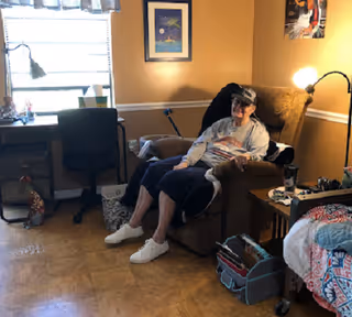 A person sitting in a recliner in a cozy private room with a bed, desk, and lamp by a window.