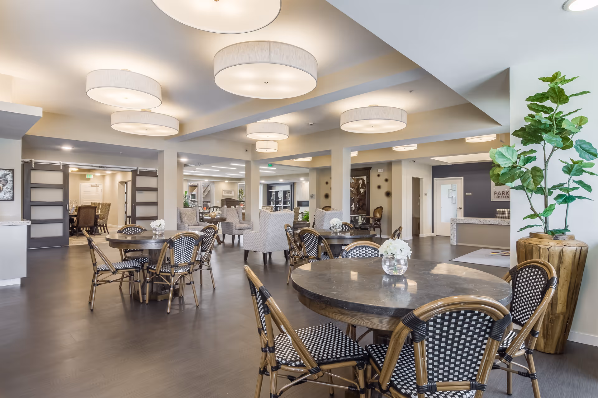 A spacious and well-lit common area in a retirement facility featuring multiple round tables with chairs, comfortable armchairs, modern ceiling lights, and decorative plants. The room has a neutral color palette with light walls and dark flooring, and there is a reception desk visible in the background with a sign that reads 'Park Creek Active Retirement'.
