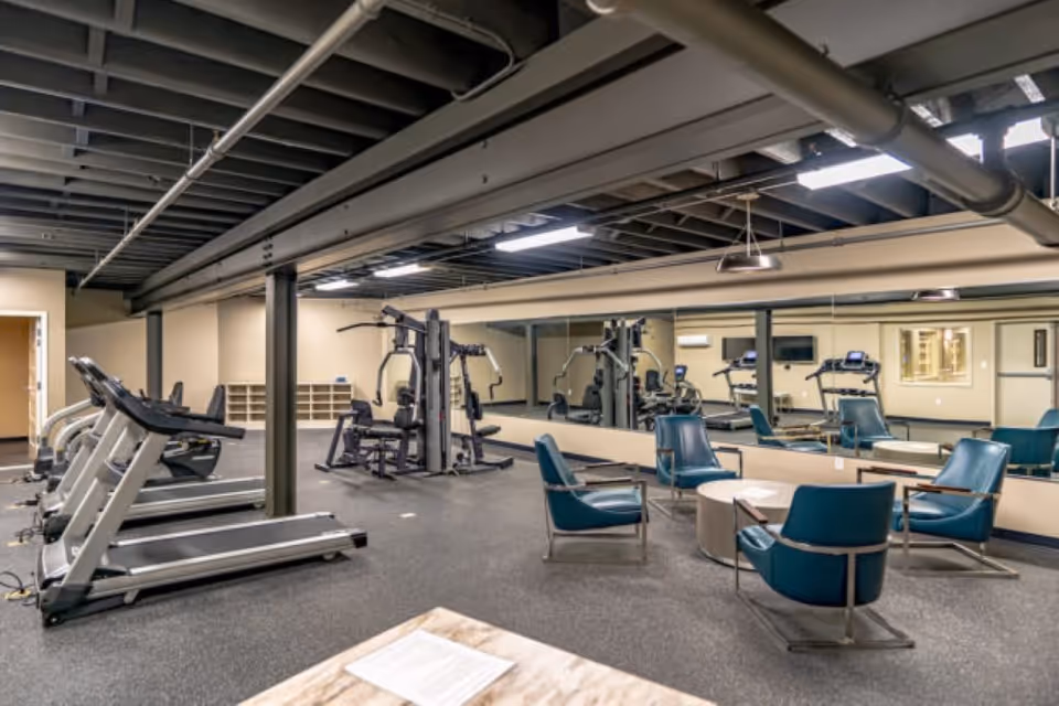 Interior view of a fitness room in a senior living facility featuring treadmills, weight machines, and a seating area with blue chairs and a round table. The room has a large mirror on one wall and exposed ceiling beams with lighting fixtures.