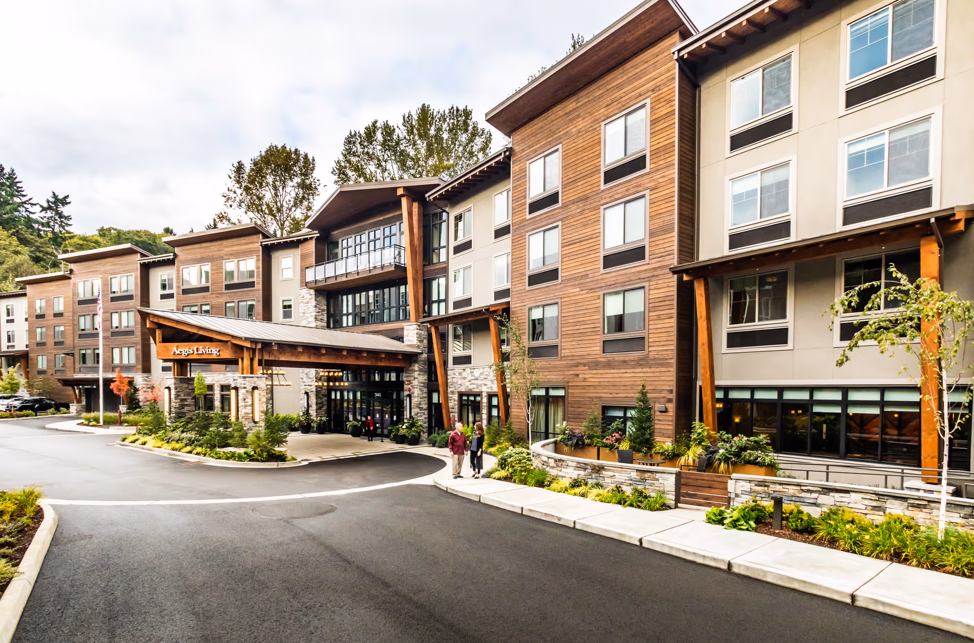 Exterior view of Aegis Living Mercer Island, a modern multi-story senior living facility with wood and stone accents. The entrance features a covered drop-off area with landscaping and a curved driveway. Two people are walking on the sidewalk near the entrance.