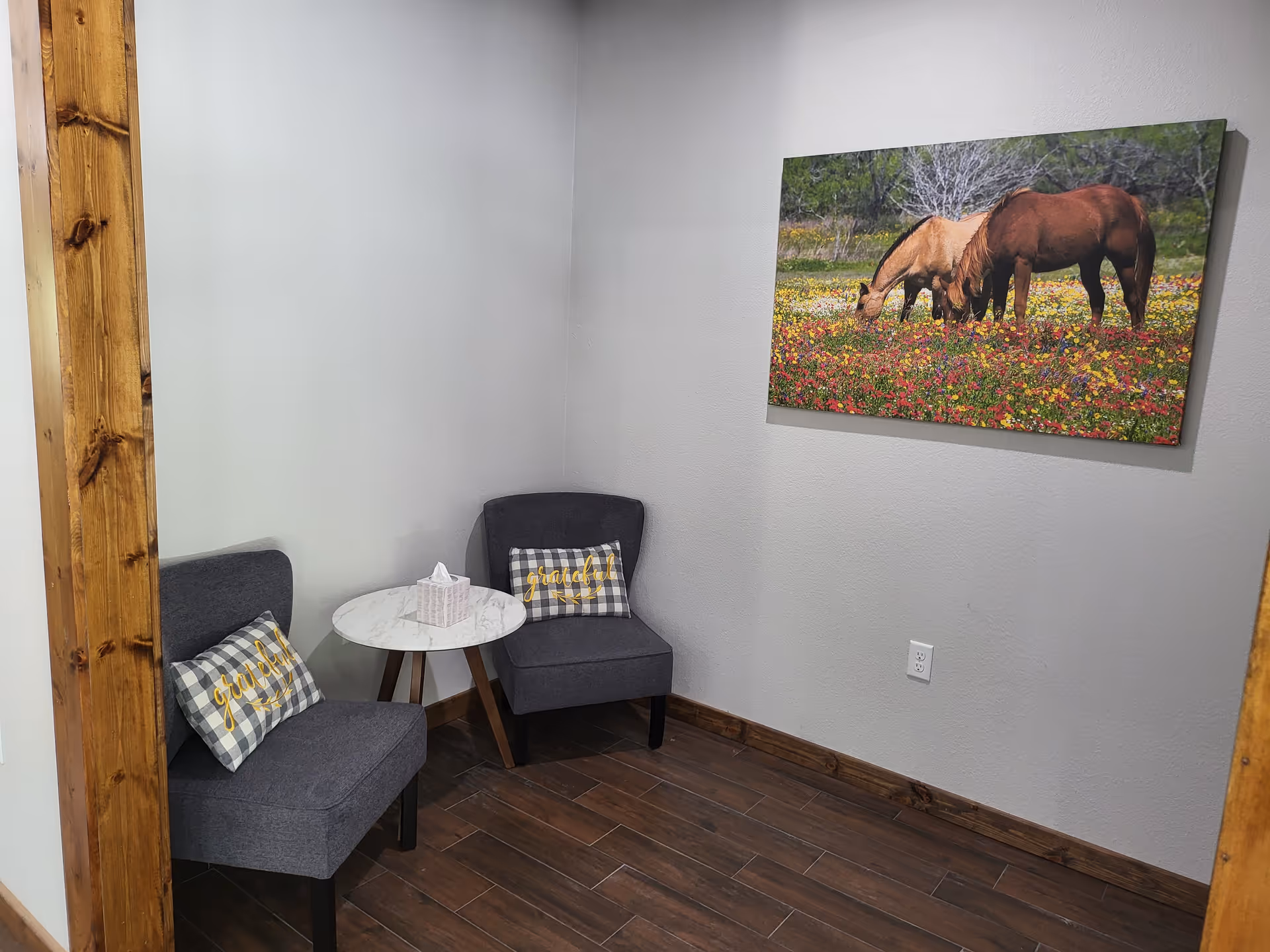 Small seating area with two gray chairs, a round side table with a tissue box, and a horse painting on the wall.