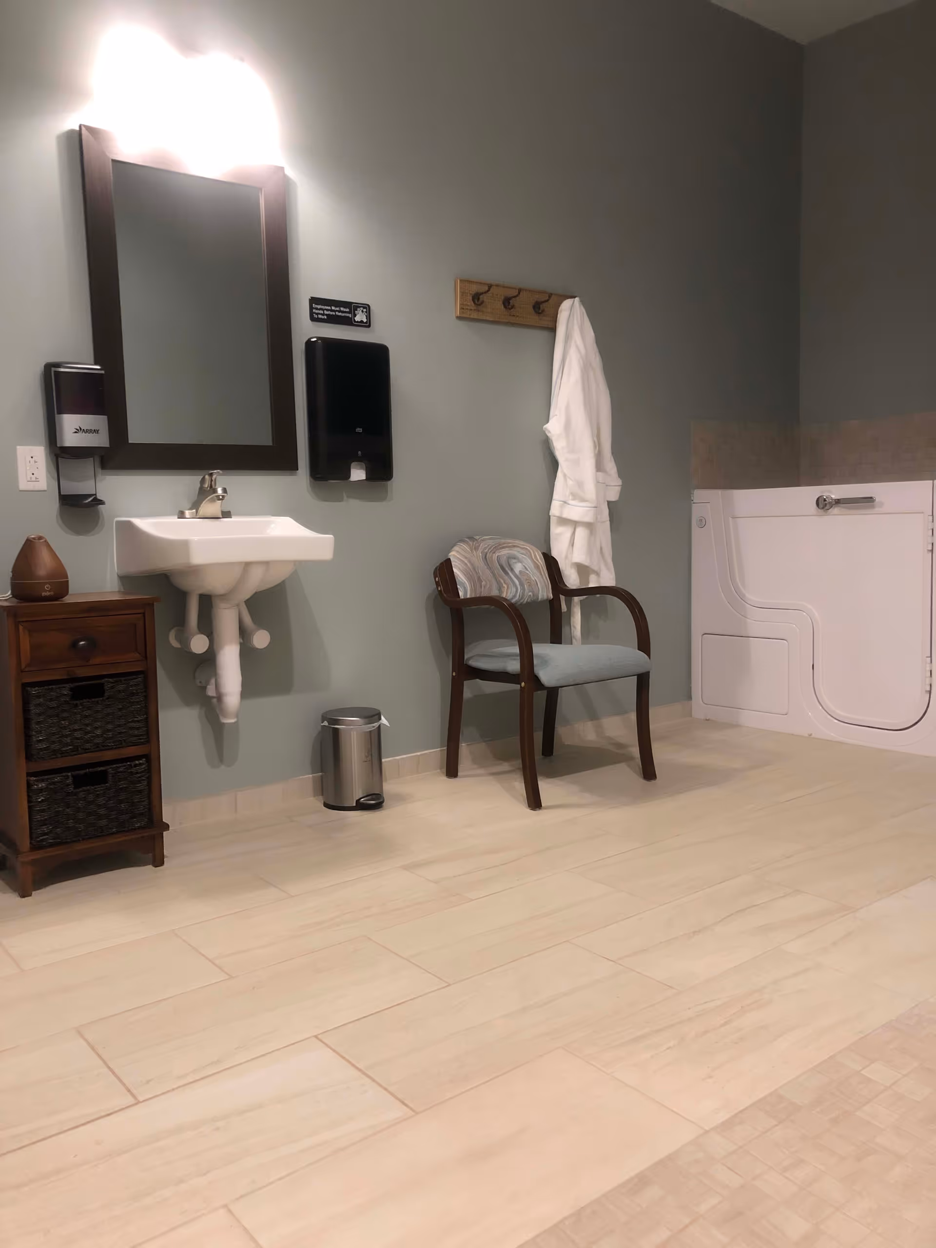 Accessible bathroom with a wall-mounted sink and mirror, a small cabinet and chair, robe on hooks, and a white walk-in tub.