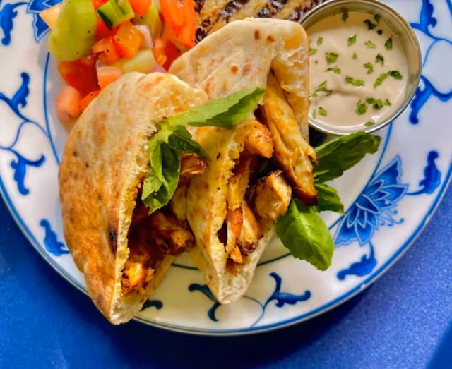 Plate with two pita sandwiches filled with grilled chicken and fresh greens, served with a side of creamy dipping sauce and a colorful cucumber and tomato salad on a blue and white patterned plate.