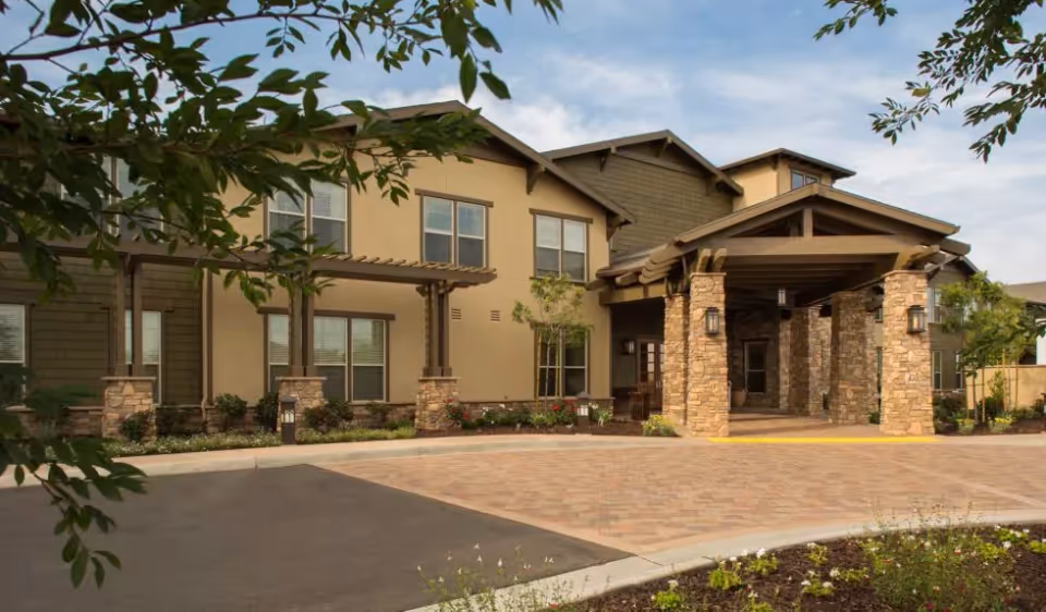 Exterior view of a senior living facility building with a covered entrance supported by stone pillars, beige and brown walls, multiple windows, and landscaped greenery including trees and flower beds.