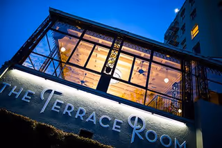 Exterior view of The Terrace Room building at dusk, showing large windows with warm interior lighting and decorative ironwork.