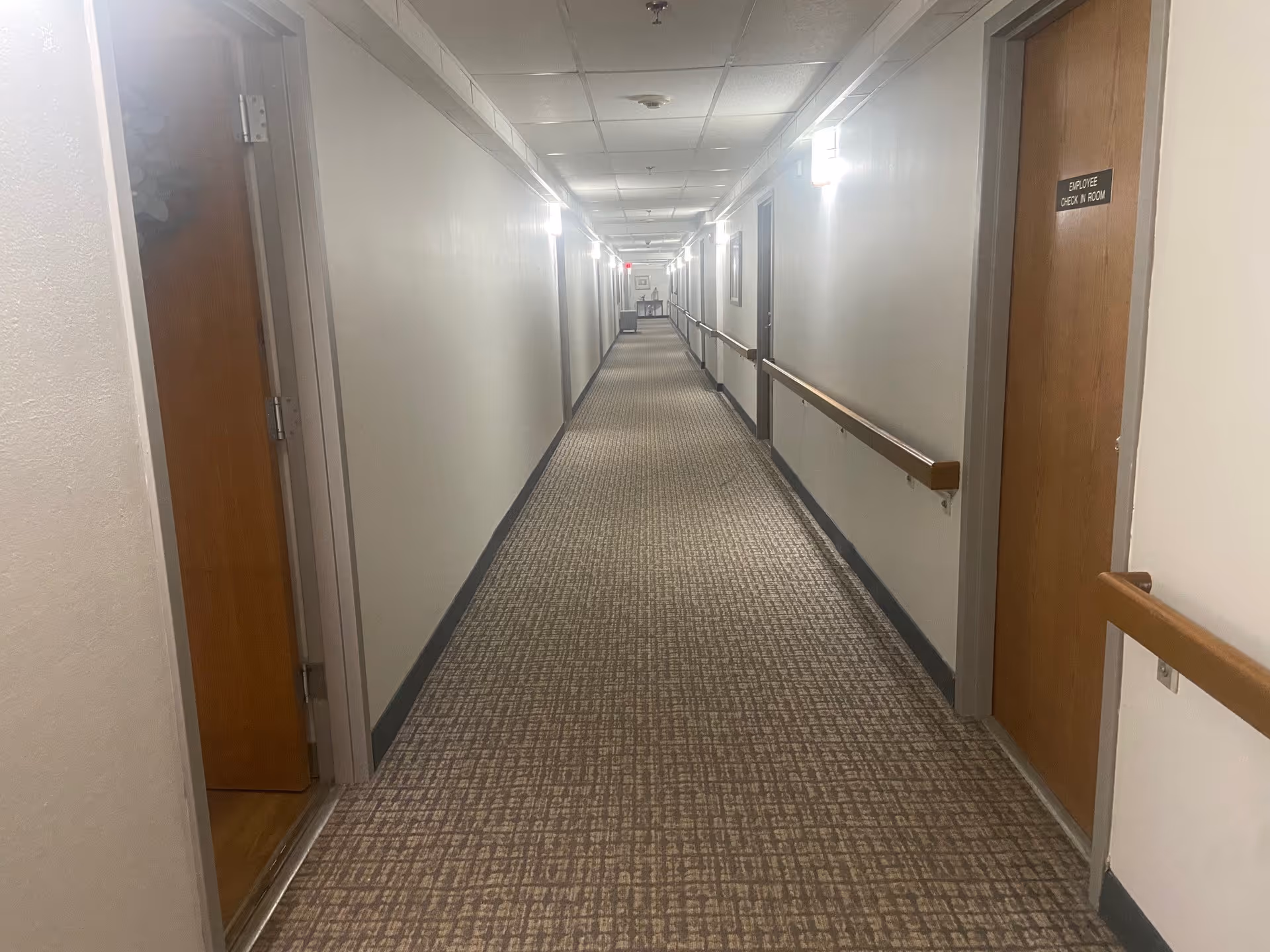 Long carpeted interior hallway with handrails and closed wooden doors on both sides in a senior living facility.