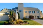 Exterior view of a two-story senior living facility building with beige walls and a gray roof, featuring a covered entrance and a statue surrounded by landscaping in the front lawn.