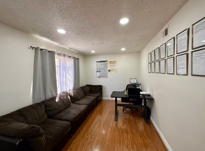A small office or waiting room with a long brown sectional sofa along one wall, a window with gray curtains, a black desk with a chair, and framed documents hanging on the opposite wall. The floor is wood laminate and the ceiling has recessed lighting.