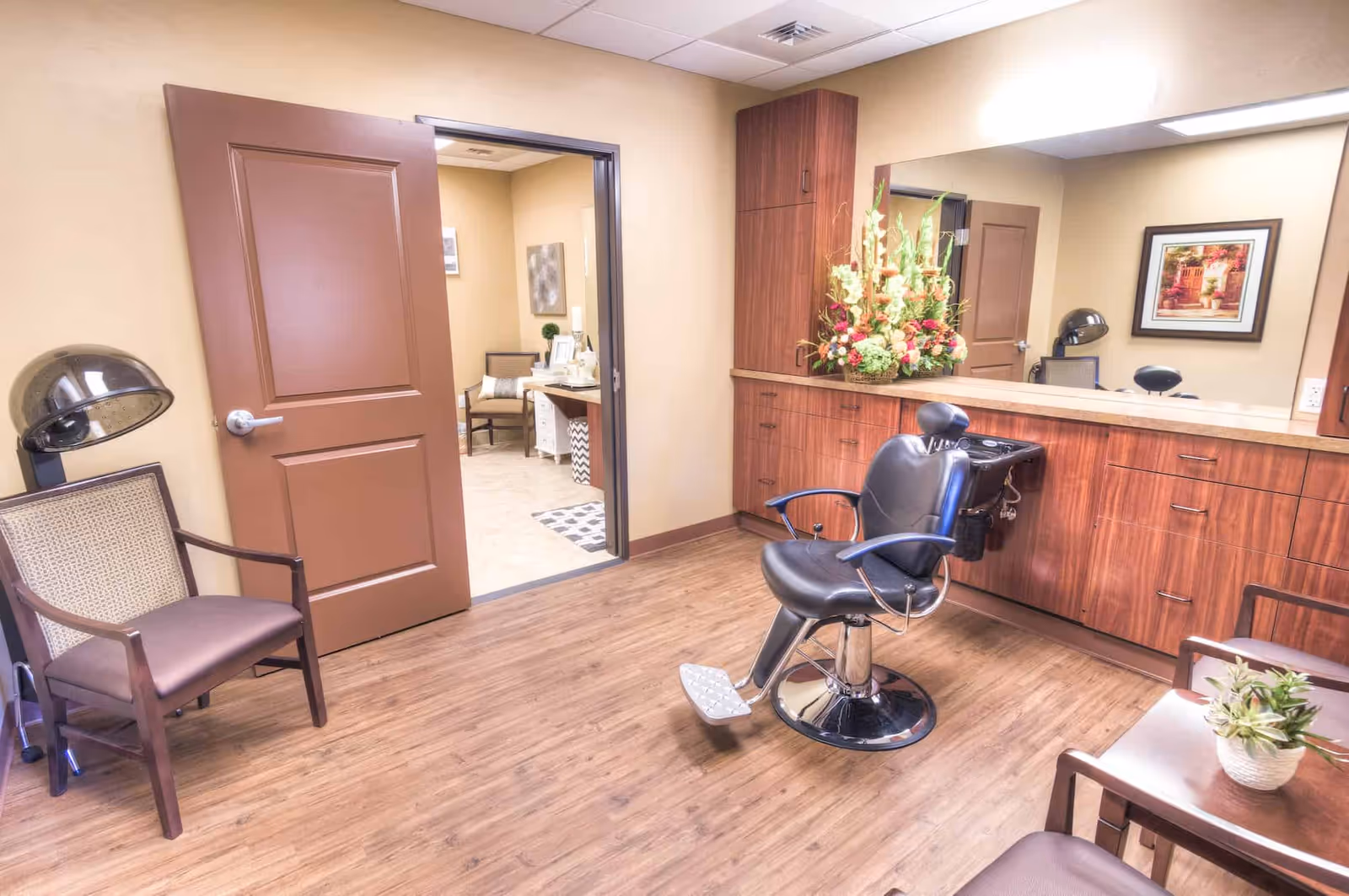 Interior view of a salon room in a memory care facility with a black salon chair in front of a large mirror and wooden cabinets. There are two brown chairs, one with a hair dryer attached, and a small table with a plant. The room has wooden flooring and beige walls, with an open door leading to another room with additional seating and decor.