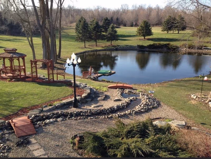 A peaceful outdoor garden area featuring a small pond surrounded by grass and trees. There is a wooden gazebo and a wooden swing set on the left side, a black lamp post with multiple lights near the center, and a winding gravel path bordered by rocks leading towards the pond. The background shows a grassy field with scattered evergreen trees and leafless deciduous trees.